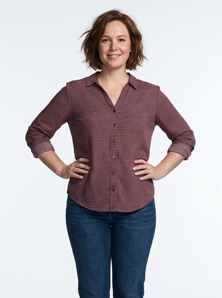 A smiling woman with short auburn hair in a burgundy shirt, hands on her hips